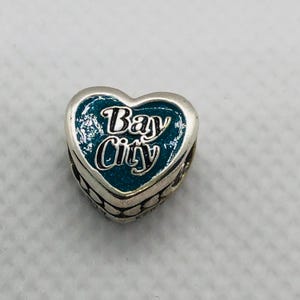 Puede incluir: Un dije de plata en forma de corazón con un esmalte de purpurina color verde azulado. El texto "Bay City" está grabado en letras plateadas en el centro del corazón. Los lados del dije muestran un patrón repetitivo de pequeños círculos.