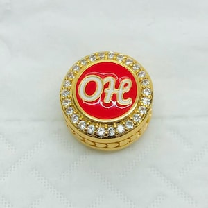 May include: A round gold-tone charm bead featuring a vibrant red enamel center with the white script letters OH. The circular face is bordered by a ring of small, clear, sparkling crystals, and the side of the bead has a textured heart pattern.