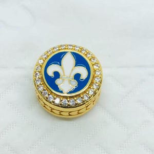 May include: A gold-colored round emblem with a white fleur-de-lis design on a blue background, surrounded by clear gemstones. The emblem has a textured edge and is set against a white background.