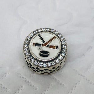 Könnte beinhalten: Ein silberner Charm-Anhänger mit einem weißen Emaille-Mittelteil, das einen Hockeypuck und gekreuzte Hockeyschläger zeigt. Der Charm ist von einem Ring aus klaren, funkelnden Kristallen umgeben. Die Seiten des Charms haben ein geometrisches Muster.