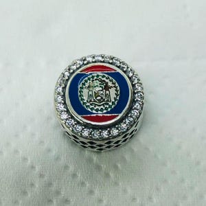 May include: A round charm featuring the flag of Belize. The charm has a blue central circle with the Belize coat of arms, framed by red. The charm is surrounded by small, clear crystals set in a silver-coloured metal.