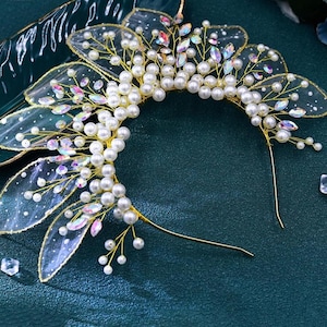 May include: A gold-toned bridal headpiece featuring a half-circle design. The headpiece is adorned with faux pearls, iridescent crystals, and delicate, sheer, petal-shaped accents. The design is set against a dark teal background.