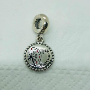 May include: A silver charm with a Ferris wheel design, featuring red accents and the word "Branson". The charm has a beaded border and a loop for attaching to a bracelet or necklace.