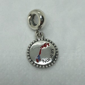 Könnte beinhalten: Ein silberner Charm-Anhänger mit einem runden Anhänger, der eine Karte von Norwegen in roter und blauer Emaille zeigt, mit dem Wort "Norway" in Schwarz. Der Charm hat einen Perlenrand und eine Schlaufe zur Befestigung.