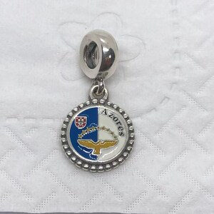 May include: A silver charm with a round pendant featuring the Azores flag design. The pendant is split into blue and white sections, with a gold bird and crown emblem. The word "Azores" is written on the pendant.