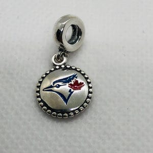 Peut inclure: Un charm en métal argenté composé d'un pendentif circulaire avec un bord perlé. Le centre présente un motif de profil d'oiseau bleu et blanc accentué par une petite feuille d'érable rouge. Le charm inclut une bélière cylindrique pour bracelet.