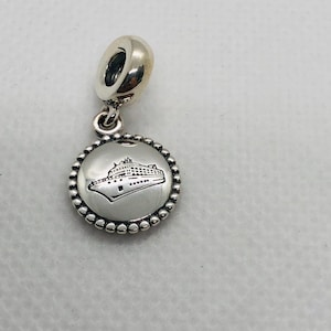 May include: A silver-toned metal charm featuring a circular pendant with a beaded edge. The centre of the pendant displays a black engraved graphic of a cruise ship. The charm includes a smooth, rounded bail at the top for attachment to a bracelet.
