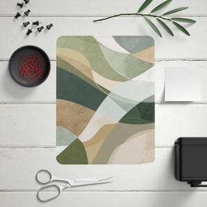 May include: An abstract art print featuring a flowing design in shades of green, beige, and white. The artwork is rectangular with rounded corners. The image is on a white wooden surface, with office supplies such as push pins, paper clips, and scissors.