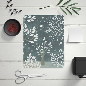 May include: A rectangular, winter-themed art print featuring white trees and falling snow on a teal background. The print is surrounded by office supplies, including push pins, paper clips, and scissors, on a white wooden surface.