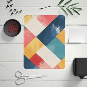 May include: Rectangular desk mat with a colourful geometric design. The pattern features overlapping brushstrokes in shades of red, blue, yellow, and white. The mat is surrounded by office supplies, including push pins, paper clips, and scissors.