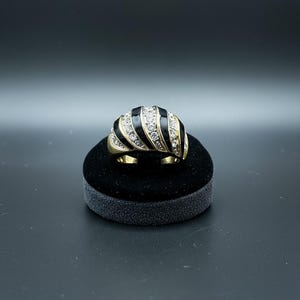 May include: A gold ring with a domed top. It has alternating black enamel and clear crystal stripes. The ring is displayed on a small black velvet stand against a dark background. The ring's design is bold and eye-catching.