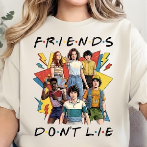 May include: Cream-colored t-shirt featuring a graphic of characters from a popular TV show. The design includes the text "FRIENDS DON'T LIE" in a playful font, with the show's logo style. The characters are depicted in a colorful, cartoon style.