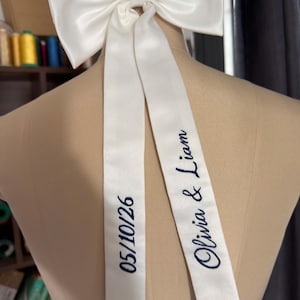May include: A large, white satin bow with long, flowing tails. The tails are embroidered with the date "05/10/26" and the names "Olivia & Liam" in navy blue script. The bow is displayed on a mannequin.