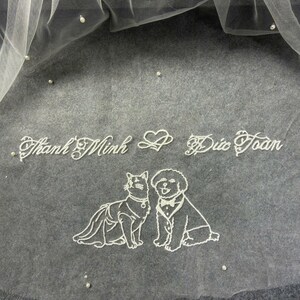 May include: A white wedding veil with scattered pearls and embroidered text. The veil features a delicate design of a cat and a dog, both in formal attire. The text reads "Thanh Minh & Duc Toan".