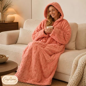 May include: A coral-colored hooded wearable blanket featuring a textured cable knit pattern. The garment has long sleeves and a loose, floor-length fit, providing full-body coverage. The fabric appears soft and plush, designed for warmth and comfort.