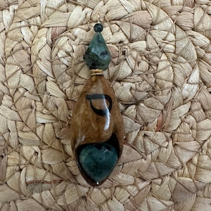 Op de afbeelding: Een handgemaakte hanger met een goudkleurige beugel. De hanger heeft een druppelvormig ontwerp met een donkerbruine en bruinachtige houtachtige uitstraling. Het heeft een donkergroene, hartvormige steen aan de onderkant en een gefacetteerde groene steen aan de bovenkant.