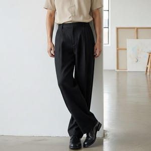 May include: A person wearing a tan short-sleeve button-down shirt and black pleated trousers. The person is wearing black dress shoes. The background is a white wall and a light-colored floor.