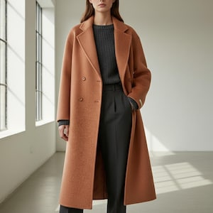 Op de afbeelding: Een lange, dubbelborstelige, roestkleurige jas. De jas heeft een klassieke revers, twee rijen knopen en een getailleerde pasvorm. Het model draagt een grijze trui en een donkergrijze broek. De jas is gemaakt van een zachte, getextureerde stof.