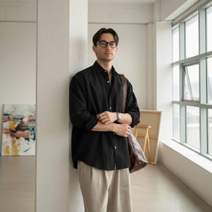 May include: A black button-down shirt paired with beige pants. The shirt is worn with the sleeves rolled up. A brown leather bag is slung over the shoulder. The person is wearing glasses and a watch.