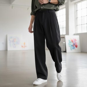 Peut inclure: Pantalon noir &agrave; jambes larges avec des plis. Le pantalon est associ&eacute; &agrave; des baskets blanches et &agrave; une chemise verte boutonn&eacute;e. Le mod&egrave;le se tient dans un espace lumineux et ouvert avec des &oelig;uvres d'art.