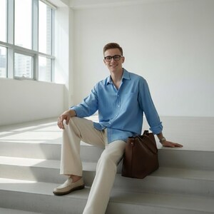 May include: A light blue button-down shirt and cream-colored pants. A brown leather tote bag sits on the steps. The person is wearing glasses and a watch.