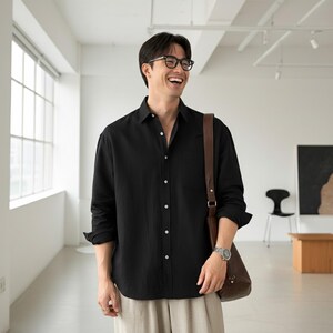 May include: Black button-down shirt with long sleeves. The shirt has a single chest pocket and is paired with light-colored pants. A brown leather shoulder bag and a silver watch are also visible.