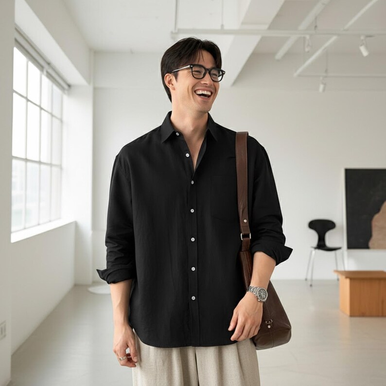 May include: Black button-down shirt with long sleeves. The shirt has a single chest pocket and is paired with light-colored pants. A brown leather shoulder bag and a silver watch are also visible.