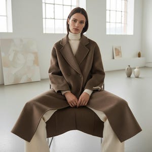 May include: A brown double-breasted coat with a wide lapel and a long, flowing design. The coat is worn over a cream-colored turtleneck and pants. The model is seated in a chair in a bright, minimalist room.
