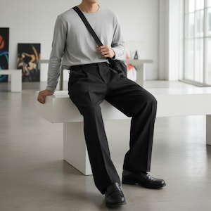 Peut inclure: Une personne portant un t-shirt gris &agrave; manches longues et un pantalon noir, assise sur un banc blanc. La personne porte des mocassins noirs et un sac en bandouli&egrave;re noir. Le cadre semble &ecirc;tre une galerie d'art moderne.
