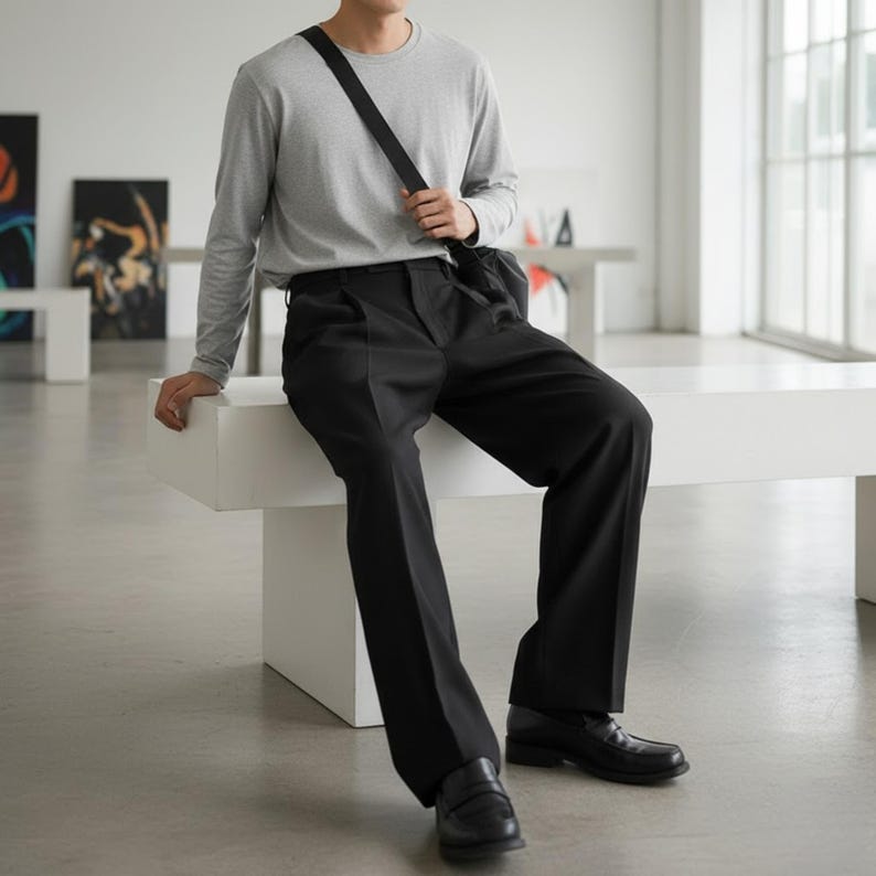 Peut inclure: Une personne portant un t-shirt gris &agrave; manches longues et un pantalon noir, assise sur un banc blanc. La personne porte des mocassins noirs et un sac en bandouli&egrave;re noir. Le cadre semble &ecirc;tre une galerie d'art moderne.
