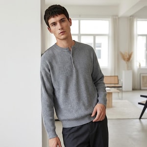May include: A gray Henley sweater with a ribbed texture. The long-sleeved shirt features a crew neck and a three-button placket. The model is wearing dark gray pants.