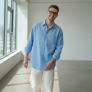 May include: A light blue button-down shirt with a chest pocket and rolled-up sleeves. The shirt is paired with cream-colored pants. The person is wearing glasses and a watch.