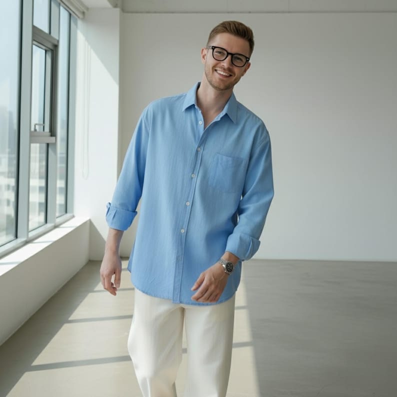 May include: A light blue button-down shirt with a chest pocket and rolled-up sleeves. The shirt is paired with cream-colored pants. The person is wearing glasses and a watch.