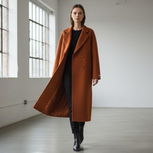 May include: A long, rust-colored coat with a double-breasted design. The coat has a classic lapel and falls to the ankles. The model is wearing a black top and pants with black boots. The coat is made of a textured fabric.