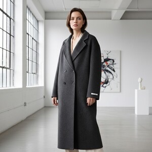 May include: A charcoal gray double-breasted coat with a classic lapel collar. The long coat features two rows of buttons and a small white label on the sleeve. The coat is worn by a person in a bright, minimalist room.