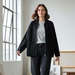 May include: A black jacket with a zipper closure and two front pockets. The jacket is worn over a gray t-shirt and paired with black jeans. The outfit is set against a backdrop of a large window and a neutral-toned wall.
