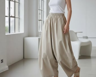 May include: A person is wearing a white tank top and beige wide-leg pants. The pants have an elastic waistband and a relaxed fit. The person is also wearing beige sandals. The background is a bright, minimalist interior with large windows.