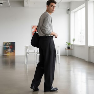 Peut inclure: Une personne portant un t-shirt gris &agrave; manches longues et un pantalon noir pliss&eacute;. Il porte un sac fourre-tout noir sur l'&eacute;paule. La personne porte des chaussures noires et marche dans un espace lumineux et ouvert.