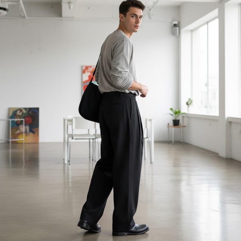 Peut inclure: Une personne portant un t-shirt gris &agrave; manches longues et un pantalon noir pliss&eacute;. Il porte un sac fourre-tout noir sur l'&eacute;paule. La personne porte des chaussures noires et marche dans un espace lumineux et ouvert.