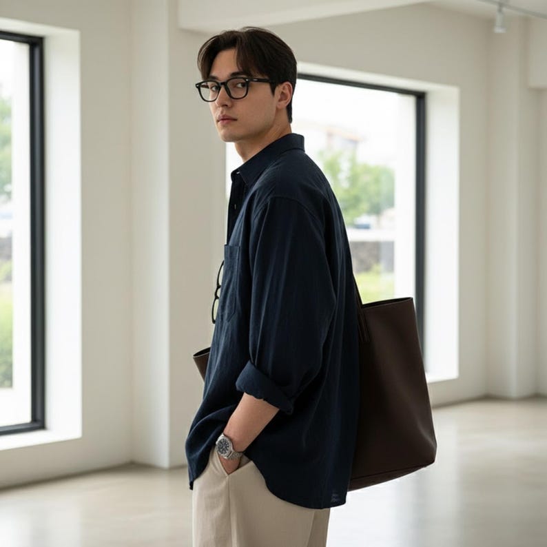 May include: A navy blue button-down shirt with rolled-up sleeves, paired with beige pants. The outfit includes a brown tote bag and a silver wristwatch. The person is wearing black-framed glasses.