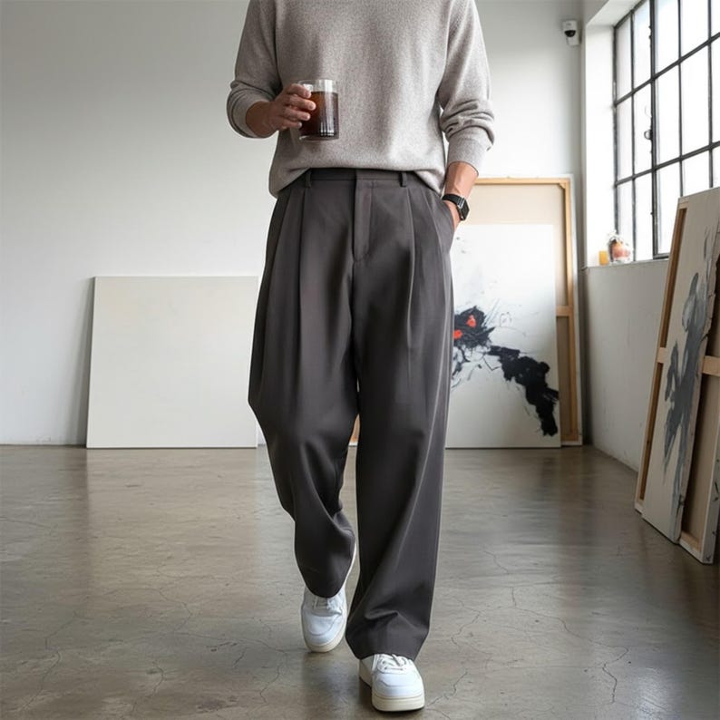 Peut inclure: Une personne portant un pull gris clair et un pantalon pliss&eacute; gris fonc&eacute;. La personne porte des baskets blanches et tient un verre d'une boisson fonc&eacute;e. Le pantalon a une coupe large.