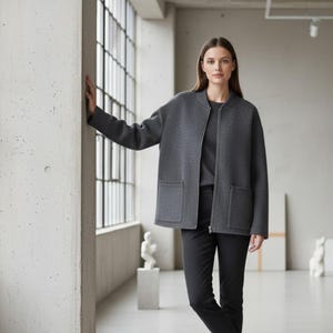 May include: A woman wearing a dark gray zip-up jacket with two front pockets, a matching gray top, and black pants. The jacket has a simple, modern design. The background features a large window and minimalist decor.