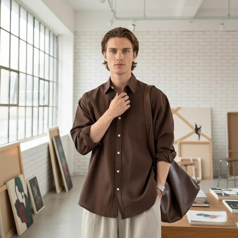 May include: A brown button-down shirt with a chest pocket and long sleeves. The shirt is worn with light-colored pants and a brown leather bag. The person is standing in a room with artwork and large windows.