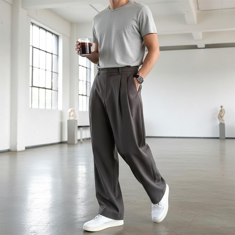 Peut inclure: Une personne portant un t-shirt gris, un pantalon pliss&eacute; gris fonc&eacute;, des baskets blanches et une montre noire. La personne tient un verre de liquide fonc&eacute;. Le cadre est un espace lumineux et minimaliste avec de grandes fen&ecirc;tres.