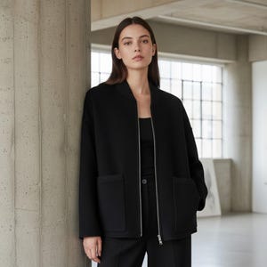 May include: A black bomber jacket with a silver zipper and two front pockets. The jacket is worn over a black top and black pants. The model is standing in front of a concrete pillar and a window.