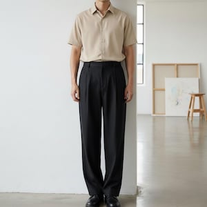 May include: A beige short-sleeve button-down shirt paired with black pleated trousers. The outfit is completed with black dress shoes. The clothing is displayed in a well-lit, minimalist setting.