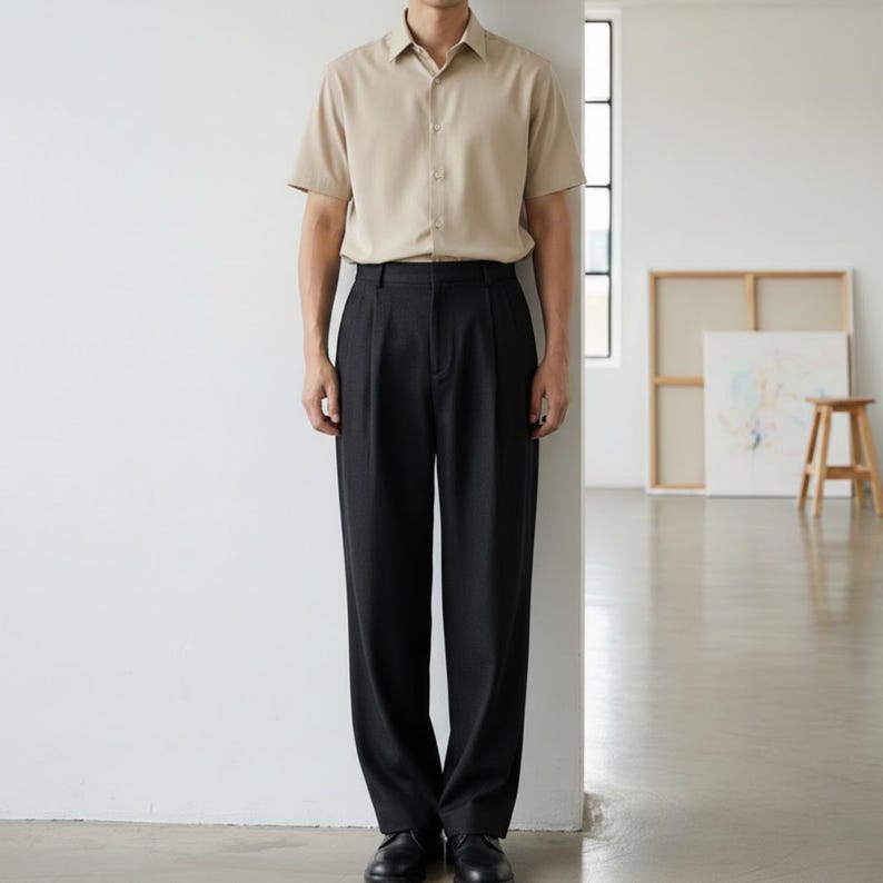 May include: A beige short-sleeve button-down shirt paired with black pleated trousers. The outfit is completed with black dress shoes. The clothing is displayed in a well-lit, minimalist setting.