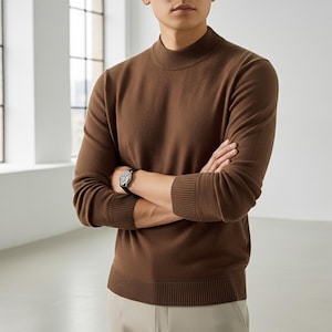 Peut inclure: Un pull marron à col montant avec des poignets et un ourlet côtelés. Le pull est de couleur unie et a des manches longues. Une montre-bracelet avec un bracelet noir est visible.