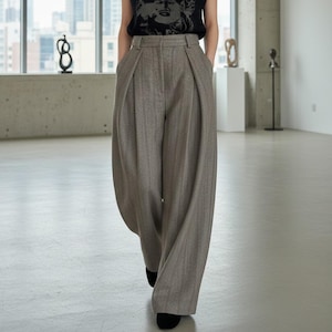 May include: Wide-leg, high-waisted trousers in a light brown herringbone pattern. The pants feature pleats at the waist and pockets. The model is wearing a black top. The pants are made of a woven fabric.