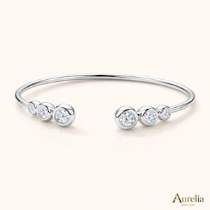 May include: A silver-toned open cuff bracelet featuring three bezel-set round clear stones on each end, graduating in size from the center outward. The metal band is smooth and polished. The text Aurelia SINCE 1989 is visible in the bottom right.